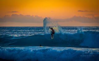 Surfer catches a wave as the sun slips behind the clouds in the sky that is glowing orange at sunset.