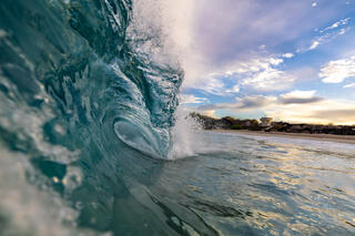 A wave curls over the top as the sunrise is seen on the shore in the distance in this wave photography image from Deron Verbeck Photography.