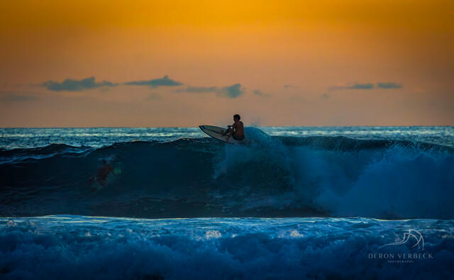 Surfer sits on his board at sunset with a orange sky and the deep blue water of the Pacific Ocean waiting for one last surf. 