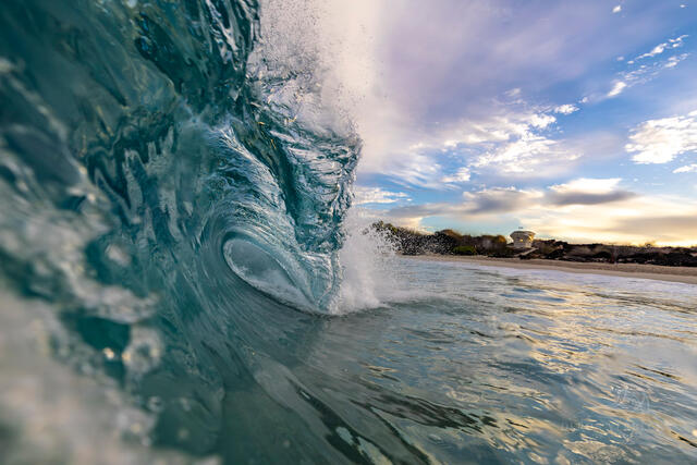 A wave curls over the top as the sunrise is seen on the shore in the distance in this wave photography image from Deron Verbeck Photography.