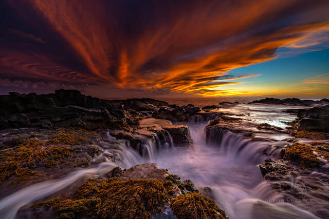 Dramatic sunset photo on the shore of Hawaii as the water rushes in and out of the old lava tube and the ocean is reflecting hues of pink & orange from the sky.